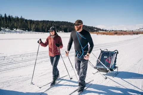 Two people cross-country ski on a snow-covered trail while pulling a child trailer.