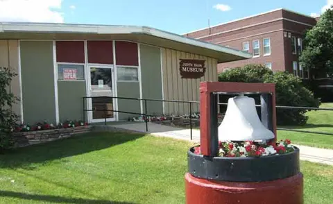 A single-story building exterior featuring a sign that reads museum and a bell display in the foreground.