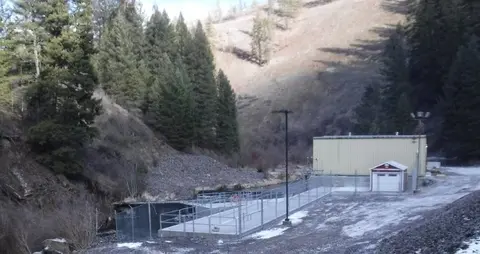 A fenced-in industrial structure and river next to a forested hillside.