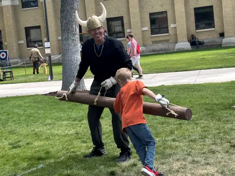 A man wearing a viking helmet and a boy carry a log together on a lawn.