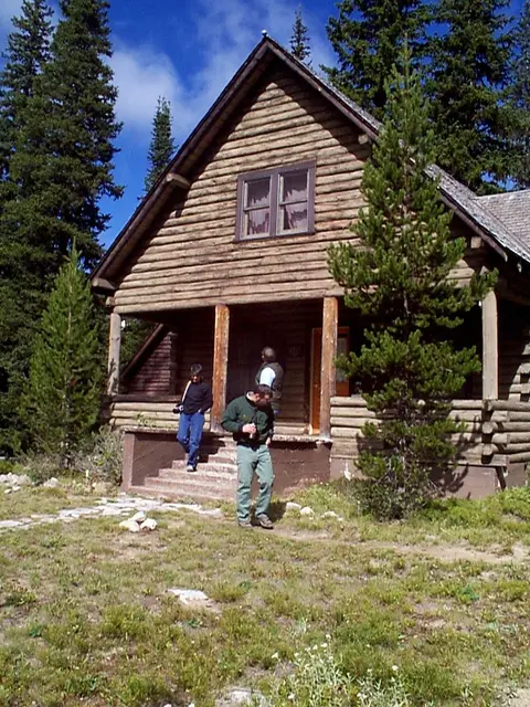 People stand outside a rustic log cabin in a wooded area.