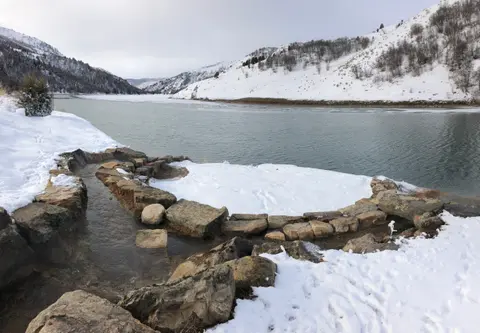 A stone-lined hot spring beside a snow-covered river and mountains.