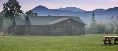 A wooden cabin stands in a grassy field in front of mountains.