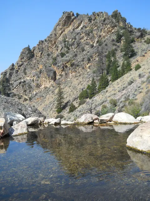 A natural pool of clear water is surrounded by rocks with a rugged mountain in the background.