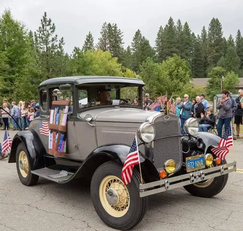 An old car decorated with American flags is driven down a street past a crowd of onlookers.