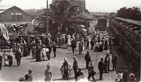 A historic black and white photograph showing a large crowd gathered near train cars and a building.