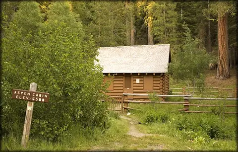 A log cabin is nestled in a forest clearing next to a trail sign.