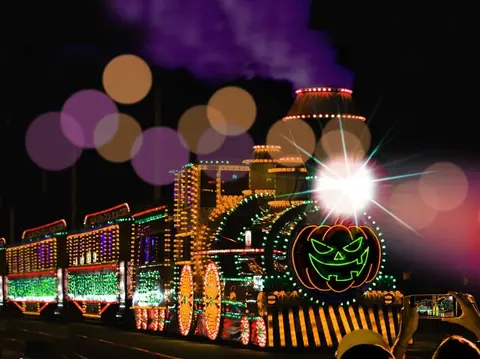 A brightly lit train decorated with lights and a jack-o'-lantern on the front travels at night.
