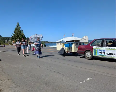 People walk in a parade behind a small float attached to a car on a road.