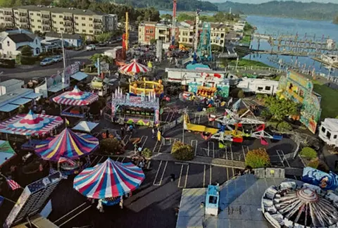 An aerial view of an outdoor carnival with rides and tents near the water.