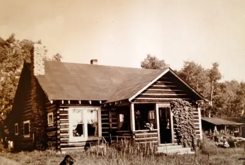 An old photograph showing a log cabin with a porch and stone chimney.