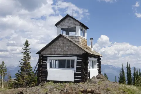 A rustic wooden lookout cabin stands on a rocky summit against a backdrop of mountains and sky.