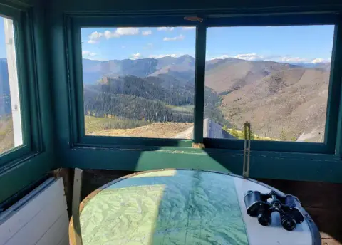 A view looking out from inside a fire lookout tower with mountains and a map visible.
