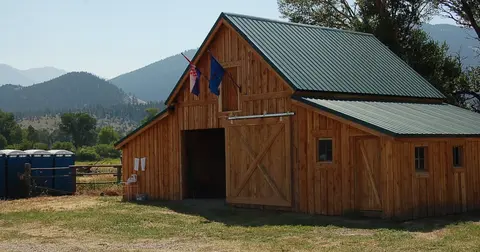 A wooden barn stands in a grassy field with mountains in the background.