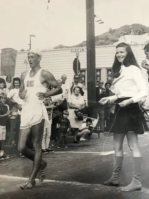 A historic black and white photo of a runner crossing a finish line with a crowd watching.