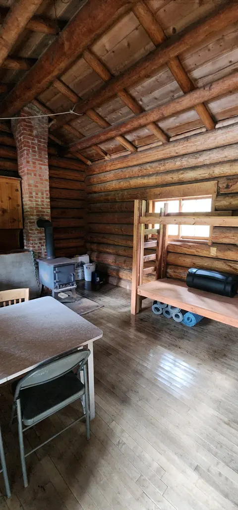 An interior view of a rustic log cabin with a wood stove and bunk beds.