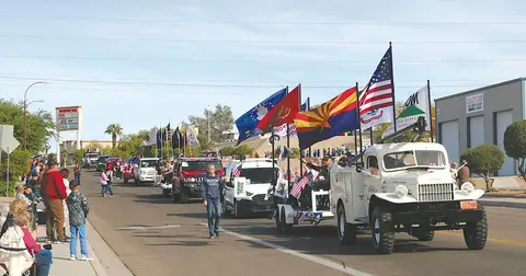 A parade with vehicles and flags travels down a street past onlookers.