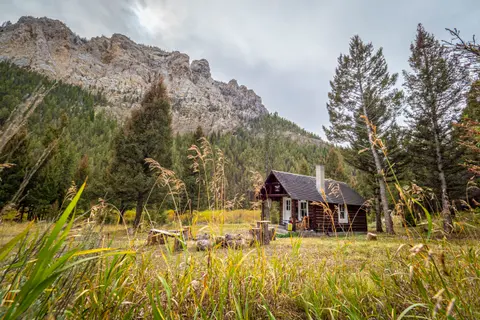 A rustic wooden cabin sits in a clearing below a steep mountain ridge.