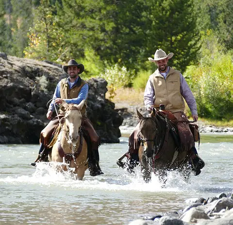 Two men ride horses through a shallow river.