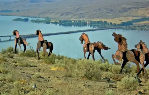 Metal horse sculptures stand on a hillside overlooking a river.