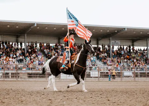 A cowgirl on horseback carries an American flag in an arena during a rodeo event.