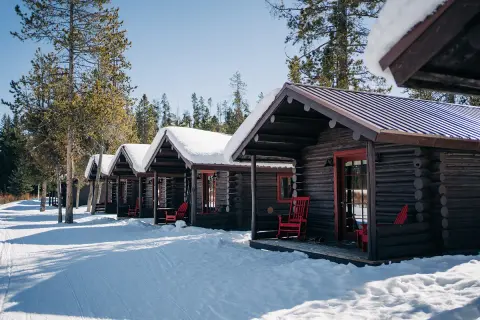 A row of rustic log cabins with snow on their roofs and red rocking chairs on their porches.