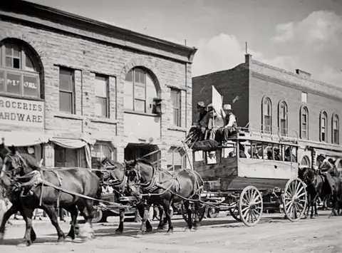 Historic black and white photo of a horse-drawn omnibus on a street in front of old brick and stone buildings.