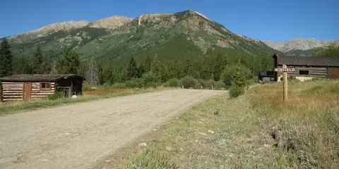 A dirt road leads through a mountain valley with wooden cabins and a large mountain in the background.