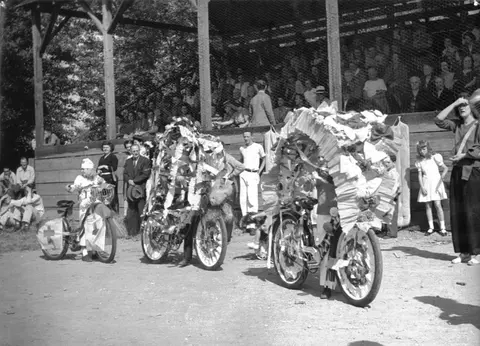 A historic black and white photo shows people with decorated bicycles in front of a grandstand.