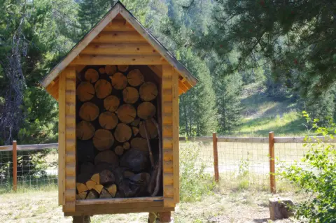 A small wooden shed filled with stacked firewood stands outdoors near a wire fence and forest.