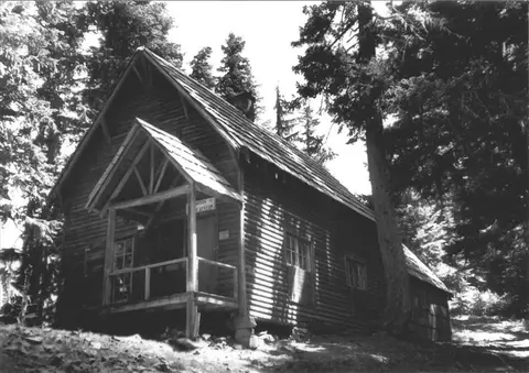 Black and white historic photo showing the exterior of a rustic wooden building surrounded by trees.