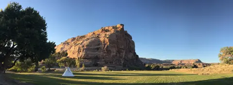 A large grassy field with a tipi sits in front of a massive rock formation under a clear blue sky.