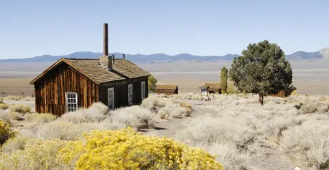 An old wooden building stands in a dry, sparse landscape with mountains in the background.