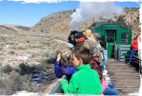 People ride on an open-air railway car through a desert landscape.