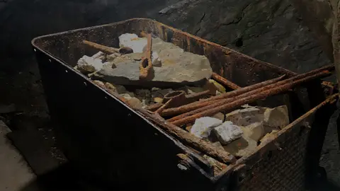 An old rusty mining cart filled with rocks and iron tools inside a dim mine.