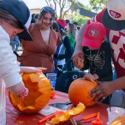 People carve pumpkins at a table outdoors.