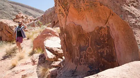 A person walks on a desert trail past rock formations with petroglyphs.