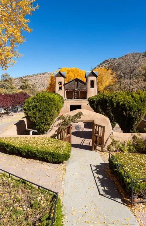 An old adobe church stands surrounded by autumn foliage and mountains under a blue sky.