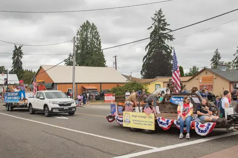 A parade featuring a white pickup truck towing a float and people riding on a decorated flatbed trailer.