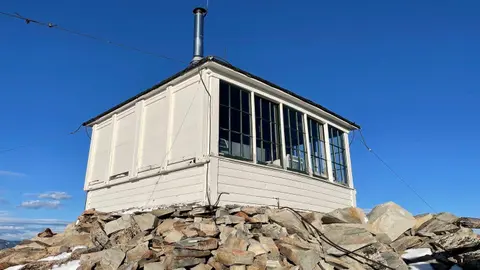 An old white lookout cabin sits atop a rocky peak against a clear blue sky.