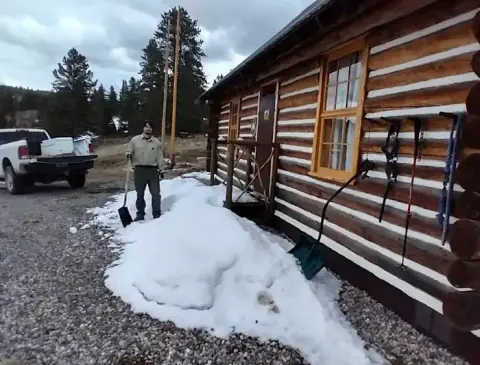A person stands with a shovel outside a log cabin with snow on the ground.