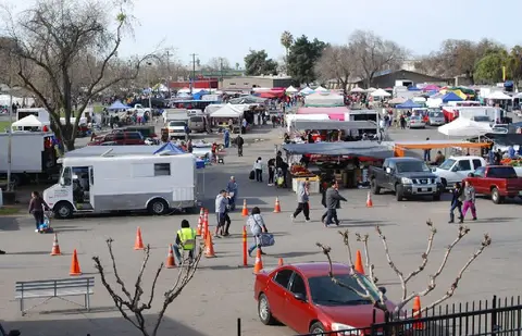 An outdoor market with many vendors, vehicles, and people walking around.