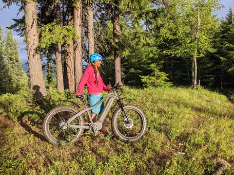 A person stands in a grassy clearing next to a mountain bike among tall trees.