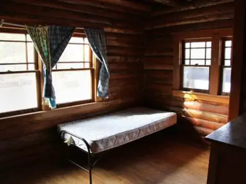 A simple cot sits inside a room with log walls and windows.