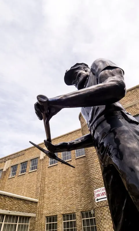 Low-angle photograph of a bronze miner statue holding a pickaxe in front of a brick building.