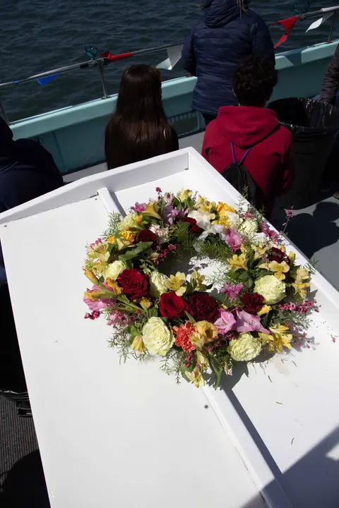 A floral wreath rests on a white surface aboard a boat with people nearby.
