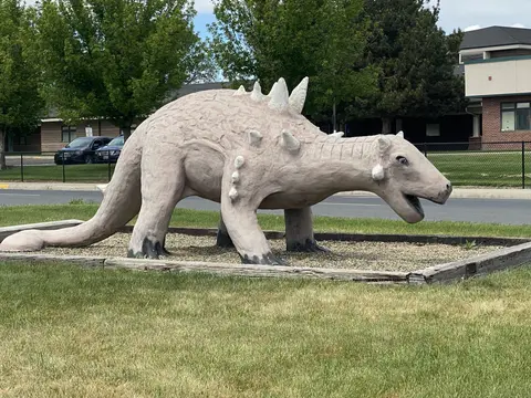 A dinosaur statue stands outdoors on a patch of grass near a street.