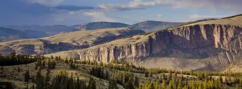 A wide landscape view of rocky cliffs, rolling hills, and a forest of evergreen trees under a cloudy sky.