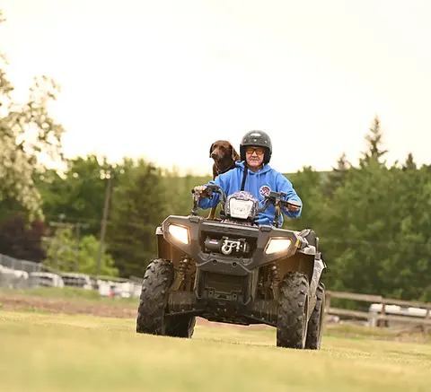 A person rides an ATV with a dog standing behind them.