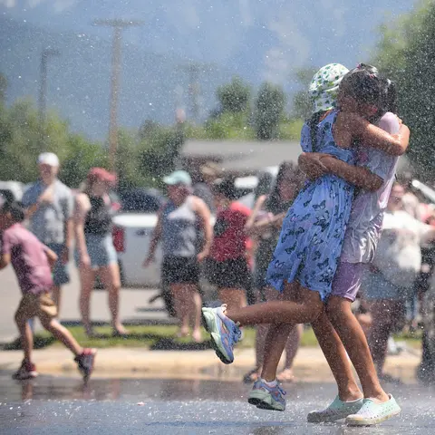 Two children hug each other in a spray of water outdoors.